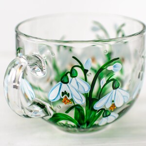 May include: A clear glass teacup featuring hand-painted snowdrop flowers with white petals, green stems, and orange centers. The design wraps around the exterior of the cup, which includes a curved handle and a wide, circular rim for drinking.