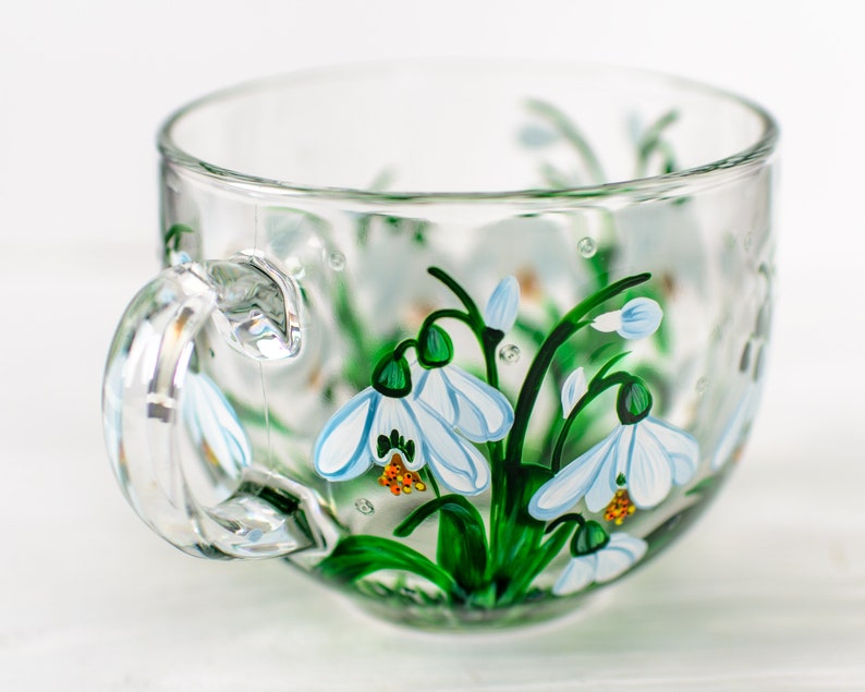 May include: A clear glass teacup featuring hand-painted snowdrop flowers with white petals, green stems, and orange centers. The design wraps around the exterior of the cup, which includes a curved handle and a wide, circular rim for drinking.