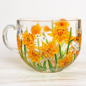 May include: A clear glass teacup with a handle, hand-painted with vibrant yellow and orange daffodils and green stems. White and gray accents are also painted on the cup. The cup sits on a white wooden surface.