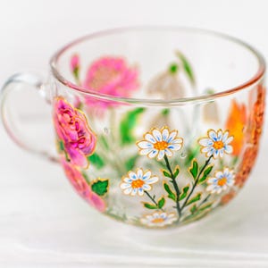 K&ouml;nnte beinhalten: Eine transparente Teetasse mit Henkel, handbemalt mit farbenfrohen Blumenmustern. Die Blumen umfassen rosa Rosen, wei&szlig;e G&auml;nsebl&uuml;mchen und orangefarbene Bl&uuml;ten, akzentuiert mit goldenen Verzierungen. Die Tasse steht vor einem wei&szlig;en Hintergrund.