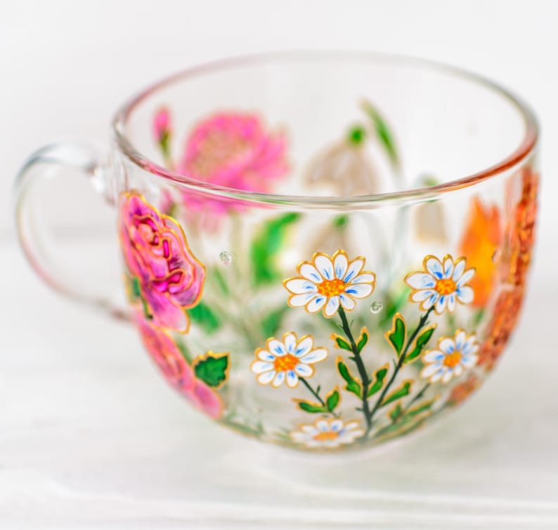 K&ouml;nnte beinhalten: Eine transparente Teetasse mit Henkel, handbemalt mit farbenfrohen Blumenmustern. Die Blumen umfassen rosa Rosen, wei&szlig;e G&auml;nsebl&uuml;mchen und orangefarbene Bl&uuml;ten, akzentuiert mit goldenen Verzierungen. Die Tasse steht vor einem wei&szlig;en Hintergrund.