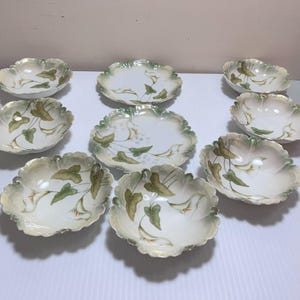 May include: A collection of nine antique porcelain dishes. The dishes have a scalloped edge with a gold trim. Each dish is decorated with a floral design featuring green leaves and white flowers. The dishes are arranged on a white surface.