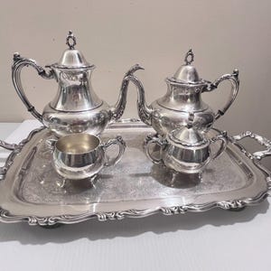 May include: A silver tea set on a silver tray. The set includes two teapots, a creamer, and a sugar bowl, all with ornate handles and lids. The tray has handles and a decorative edge.