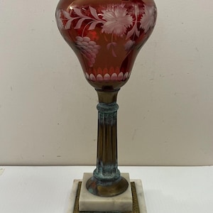 Może przedstawiać: Antyczna lampa naftowa z rubinowym kloszem ze szkła, wytrawionym we wzór kwiatowy. Lampa posiada mosiężną podstawę oraz kwadratową podstawę z marmuru i mosiądzu. Zabytkowy przedmiot dekoracyjny.