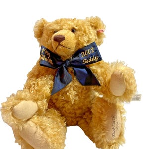 May include: A golden-brown teddy bear with a dark blue ribbon tied around its neck. The ribbon has gold text that reads "1902 Steiff" and "2002 Teddy". The bear has a tag on its ear and the text "Dick Frank 2002" on its foot.