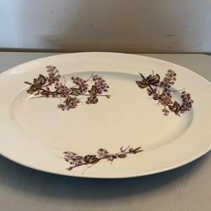 Puede incluir: Plato ovalado blanco para servir con diseño floral. El plato presenta un delicado patrón de flores moradas y hojas verdes. Los bordes están ligeramente elevados y la forma general es alargada.