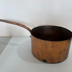 May include: A copper saucepan with a long, curved handle. The pot has a rounded body and a dark interior. The copper exterior shows signs of age and use, with a rich, warm color. This vintage cookware is ideal for cooking.