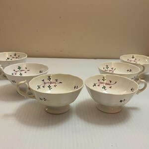 May include: Six white teacups with handles and pedestal bases are arranged on a white surface. Each cup is decorated with a floral pattern in green and purple. The cups are likely part of a tea set.