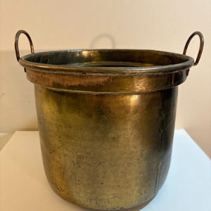 May include: A round, hammered brass bucket featuring a wide, reinforced rim and two curved metal handles attached to the sides. The surface of the vessel displays a weathered, aged patina with visible scratches and variations in the metallic tone.