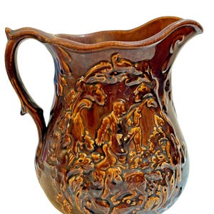 May include: A brown ceramic pitcher with a detailed relief design. The pitcher features a handle and a spout. The relief depicts a hunting scene with figures, dogs, and foliage. The pitcher has a glossy finish.