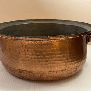 Pode incluir: Uma panela de cobre martelado com duas alças laterais. A panela tem uma forma larga e rasa com um interior escuro e um exterior martelado. O cobre tem uma cor marrom avermelhada quente.