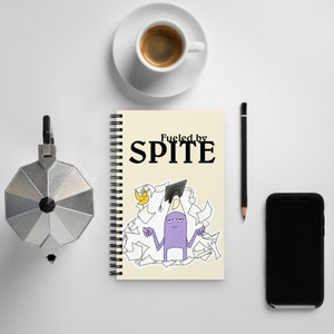 May include: A cream-coloured spiral-bound notebook with the words "Fueled by Spite" and a cartoon pencil character. A cup of coffee, a pencil, a phone, and a coffee maker are also visible. The notebook is surrounded by papers.
