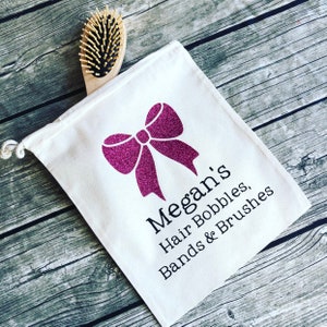 Puede incluir: Una bolsa de lona blanca con cordón de ajuste, un lazo de purpurina rosa y el texto "Megan's Hair Bobbles, Bands & Brushes".