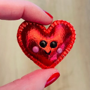 Puede incluir: Decoración de fieltro rojo en forma de corazón con una cara sonriente. El corazón tiene ojos de botón negros y mejillas rosas. Los bordes están cosidos con hilo blanco. El corazón se sostiene entre dos dedos.