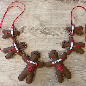 May include: A garland of six brown felt gingerbread figures connected by red ribbons. Each figure features stitched details, small red buttons, and white letter beads spelling names including Edith, Bonnie, Nanny, Diaddiad, Elizia, and Henry.