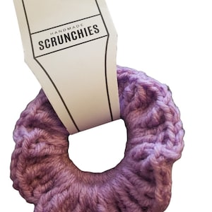 Puede incluir: Un scrunchie de color morado tejido a crochet con una etiqueta blanca que dice "Handmade Scrunchies".