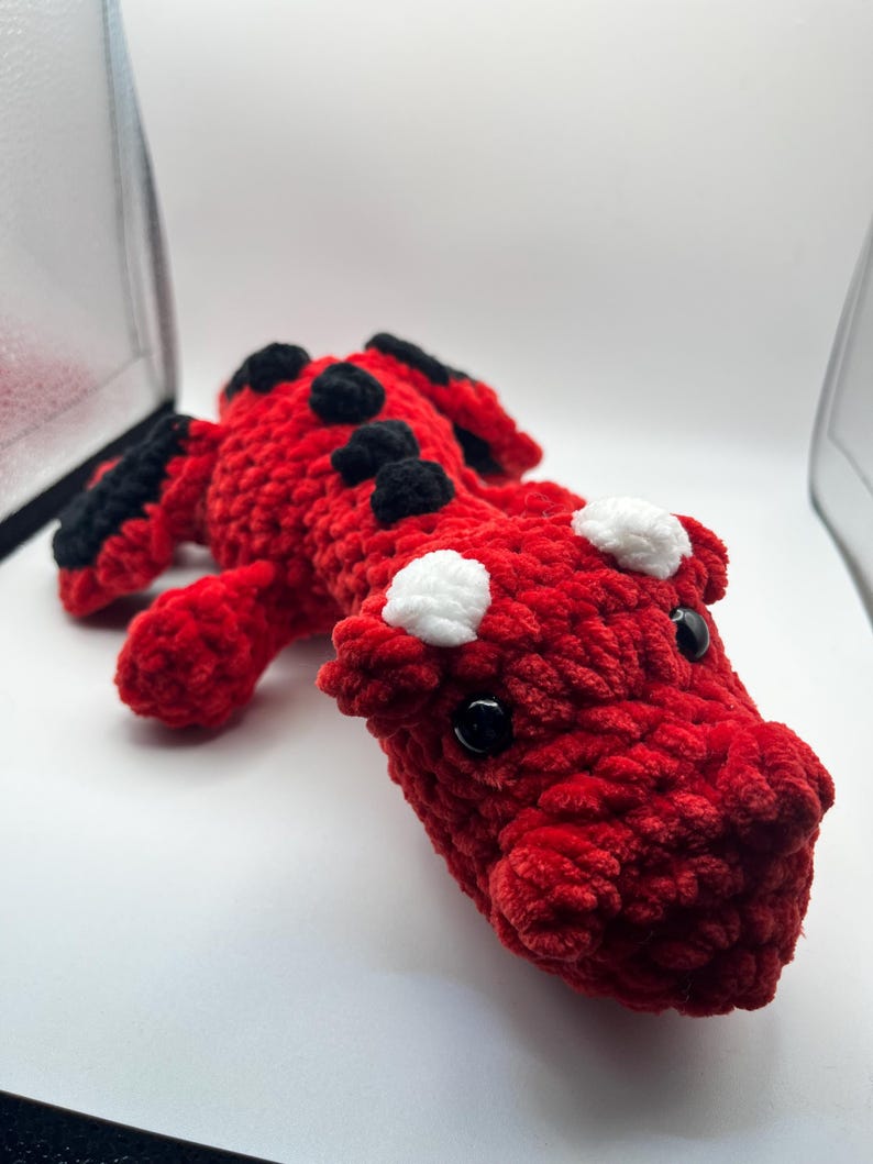 Puede incluir: Un drag&oacute;n de ganchillo rojo hecho a mano con detalles en negro y blanco. El drag&oacute;n tiene ojos negros de bot&oacute;n. El cuerpo del drag&oacute;n es rojo con manchas negras y manchas blancas en la cabeza.