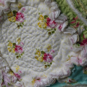 May include: Close-up of a handmade quilt with a floral pattern. The quilt features a white background with yellow polka dots and spiral stitching. Pink and yellow roses are scattered throughout. The edges are trimmed with ruffled fabric in shades of pink, white, and green.
