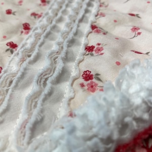 May include: Close-up of a floral fabric with a delicate pattern of small pink flowers on a cream background. White ruffled trim adds texture and detail to the design. The fabric appears soft and lightweight.