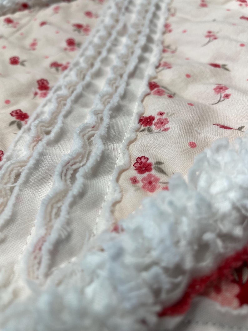 May include: Close-up of a floral fabric with a delicate pattern of small pink flowers on a cream background. White ruffled trim adds texture and detail to the design. The fabric appears soft and lightweight.