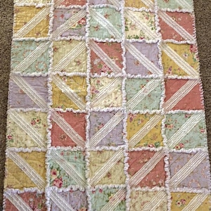 May include: A patchwork quilt with a floral pattern in pastel colors. The quilt features squares of pink, yellow, green, and lavender fabric, separated by white lace trim. The quilt is a cozy, handmade item.
