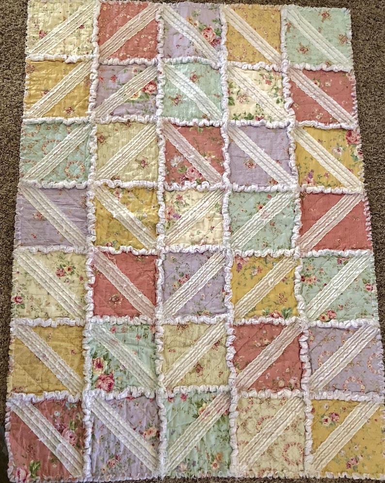 May include: A patchwork quilt with a floral pattern in pastel colors. The quilt features squares of pink, yellow, green, and lavender fabric, separated by white lace trim. The quilt is a cozy, handmade item.