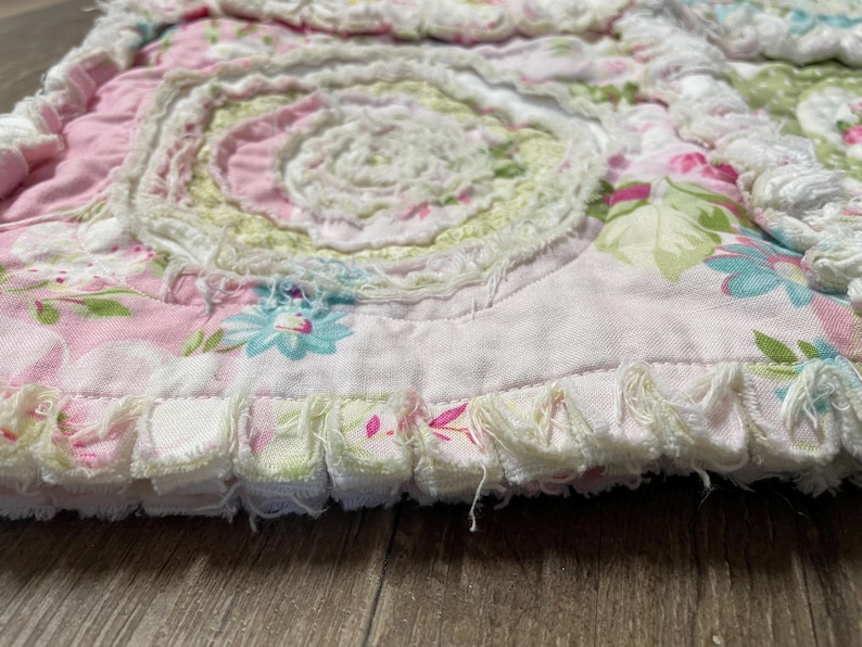 May include: A close-up of a floral patterned quilt with a spiral design. The quilt features pink, green, and white colors with a ruffled edge. The floral pattern includes blue and pink flowers.