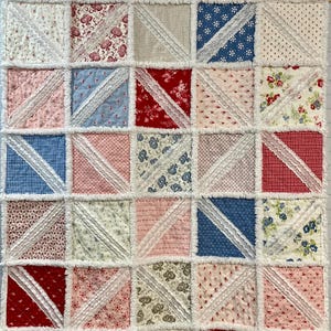 May include: A patchwork quilt with a repeating pattern of squares. Each square is divided diagonally with white lace trim. The squares feature various floral and patterned fabrics in red, blue, pink, and cream colors. The quilt has a fringed edge.