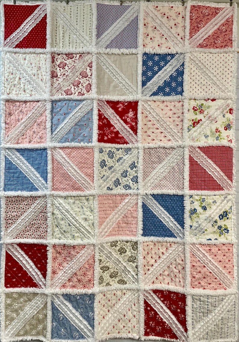 May include: A patchwork quilt with a repeating pattern of squares. Each square is divided diagonally with white lace trim. The squares feature various floral and patterned fabrics in red, blue, pink, and cream colors. The quilt has a fringed edge.