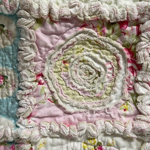 May include: Close-up of a handmade quilt with a floral pattern. The quilt features a light blue section with floral medallions, a pink section with a central rosette, and white ruffled edges. The color palette includes pink, white, green, and blue.