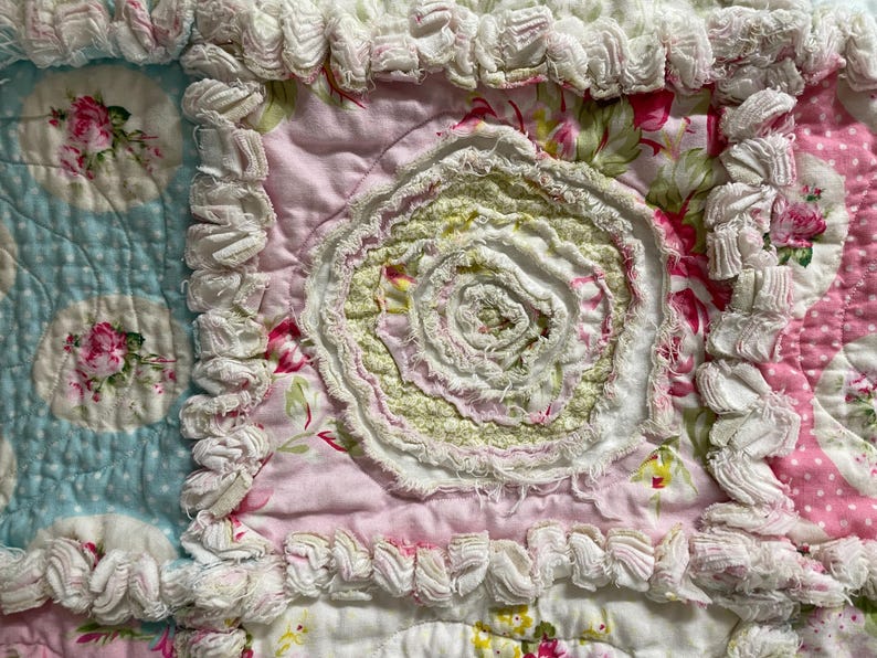 May include: Close-up of a handmade quilt with a floral pattern. The quilt features a light blue section with floral medallions, a pink section with a central rosette, and white ruffled edges. The color palette includes pink, white, green, and blue.