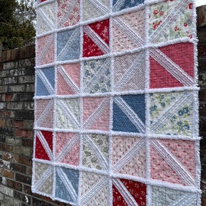 May include: A handmade quilt with a patchwork design. The quilt features a variety of fabrics in red, white, blue, and pink, arranged in a grid pattern. White, ruffled fabric separates the squares. The quilt is hanging on a brick wall.