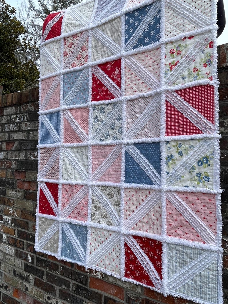 May include: A handmade quilt with a patchwork design. The quilt features a variety of fabrics in red, white, blue, and pink, arranged in a grid pattern. White, ruffled fabric separates the squares. The quilt is hanging on a brick wall.