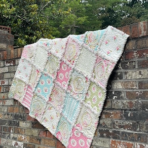 May include: A handmade patchwork quilt with a variety of floral and striped patterns in pastel colors. The quilt is made of squares of fabric, some with a ruffled texture, and is draped over a brick wall. The color palette includes pink, blue, green, and white.