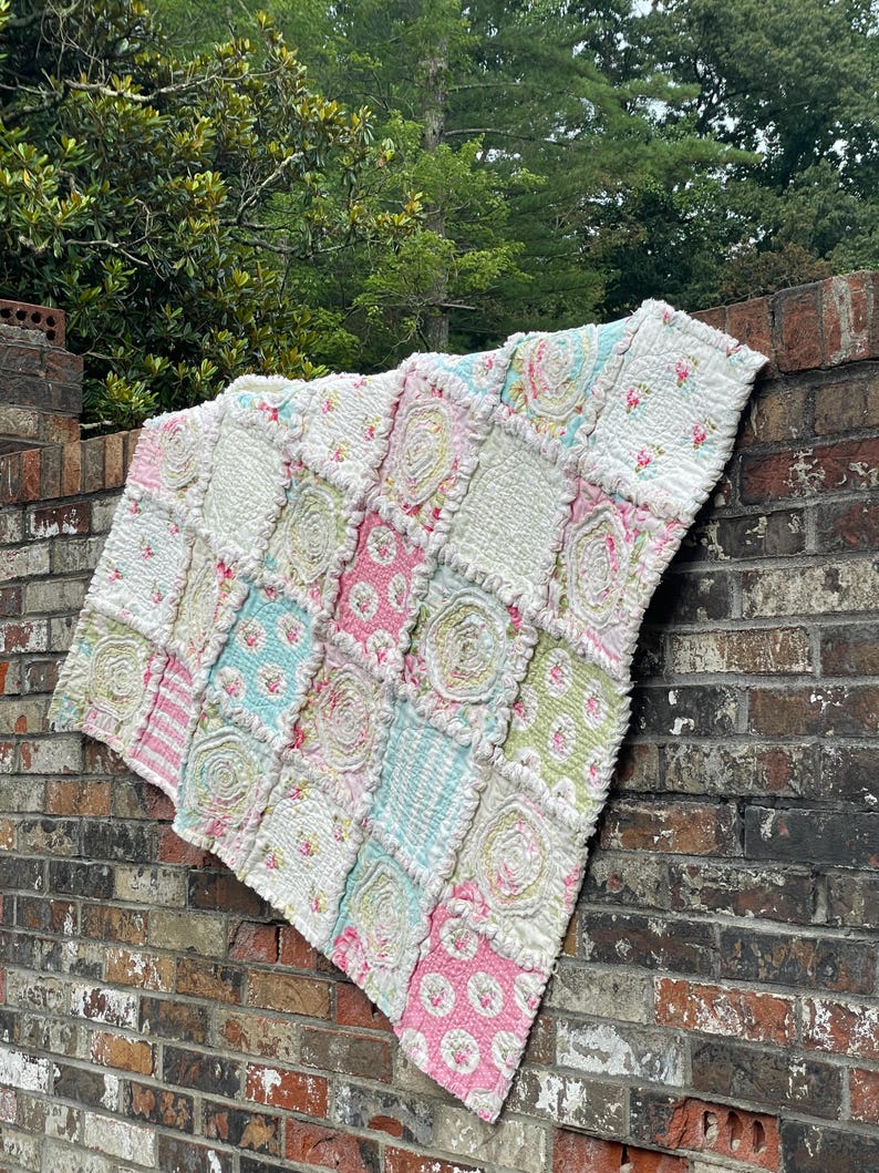 May include: A handmade patchwork quilt with a variety of floral and striped patterns in pastel colors. The quilt is made of squares of fabric, some with a ruffled texture, and is draped over a brick wall. The color palette includes pink, blue, green, and white.