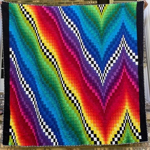 May include: A vibrant, handmade quilt with a rainbow gradient design. The quilt features a chevron pattern with black and white checkered accents. The edges are bordered with black fabric and a white dotted trim. A colorful and cozy home decor item.