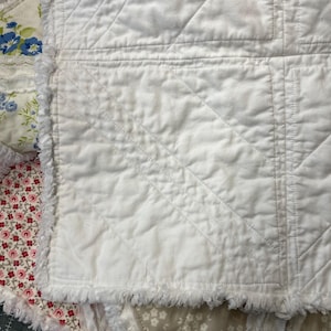 May include: Close-up of a white quilted blanket with a diamond pattern. The blanket has a fringed edge and is layered with floral patterned fabric in shades of pink, blue, and beige. The quilt is a handmade item.