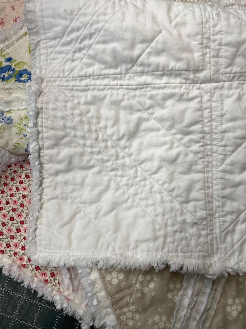 May include: Close-up of a white quilted blanket with a diamond pattern. The blanket has a fringed edge and is layered with floral patterned fabric in shades of pink, blue, and beige. The quilt is a handmade item.