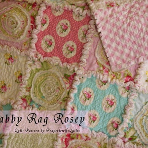 May include: A patchwork quilt with a floral pattern in shades of pink, green, and blue. The quilt features a ruffled edge and the text "Shabby Rag Rosey". The quilt is made of various fabrics with a vintage, shabby chic aesthetic.