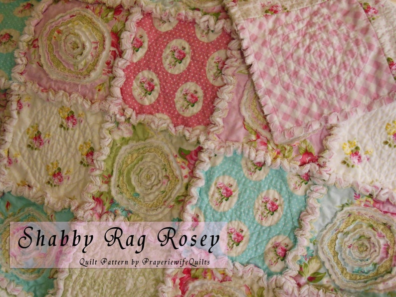 May include: A patchwork quilt with a floral pattern in shades of pink, green, and blue. The quilt features a ruffled edge and the text "Shabby Rag Rosey". The quilt is made of various fabrics with a vintage, shabby chic aesthetic.