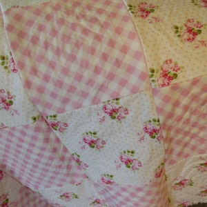 May include: A patchwork quilt featuring a mix of patterns. The quilt has sections of pink and white gingham, white fabric with pink rose bouquets, and white fabric with small pink polka dots. The quilt is a soft, cozy home decor item.