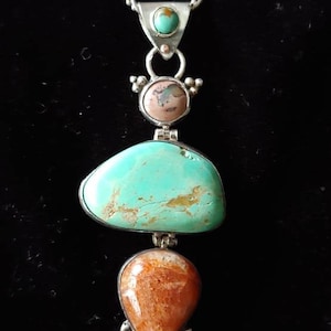 Può includere: Una collana d'argento con un ciondolo in turchese, rosa e arancio. Il ciondolo è composto da tre pietre, ciascuna con una forma e una dimensione diverse. La collana è un'ottima aggiunta a qualsiasi collezione di gioielli.