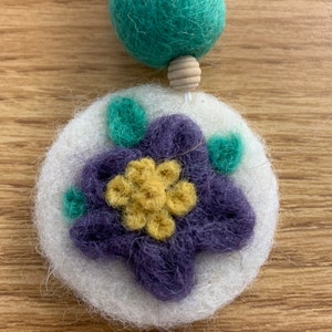 May include: A white felt circle with a purple felt flower with a yellow center and green felt leaves. The flower is surrounded by a white felt border.