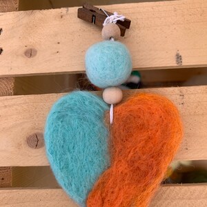 May include: A felt heart ornament with a teal and orange color scheme. The heart is strung on a white cord with wooden beads and a wooden clothespin.