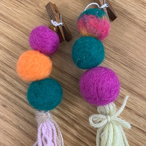 May include: Two colorful felt ball garlands with wooden clothespins at the top. Each garland has a pink, orange, and green felt ball with a tassel at the bottom. One tassel is pink and beige, and the other is beige.