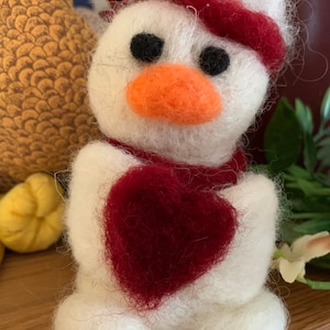Peut inclure: Un bonhomme de neige en feutre blanc avec un chapeau et une écharpe en feutre rouge, tenant un cœur en feutre rouge. Le bonhomme de neige a des yeux noirs et un nez en feutre orange.
