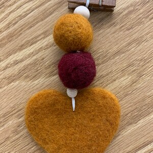 May include: A felt heart garland with a brown felt heart, a burgundy felt ball, and a mustard yellow felt ball. The garland is strung on a white string and attached to a wooden clothespin.