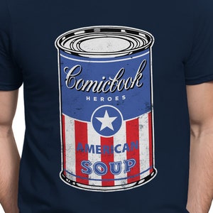 May include: A navy blue t-shirt with a vintage-style illustration of a can of soup. The can is red, white, and blue with the text "Comicbook Heroes American Soup".