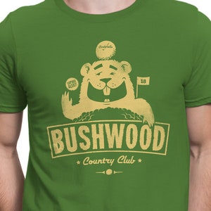 May include: A green t-shirt with a cartoon image of a bear wearing a golf hat and holding a golf club. The text "Bushwood Country Club" is printed on the shirt.