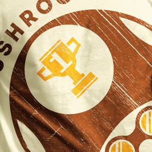 May include: Close-up of a cream-colored t-shirt with a distressed brown design. The design features the word "SHROOM" in a circular pattern, surrounding a gold trophy graphic. The shirt has a vintage, worn look.